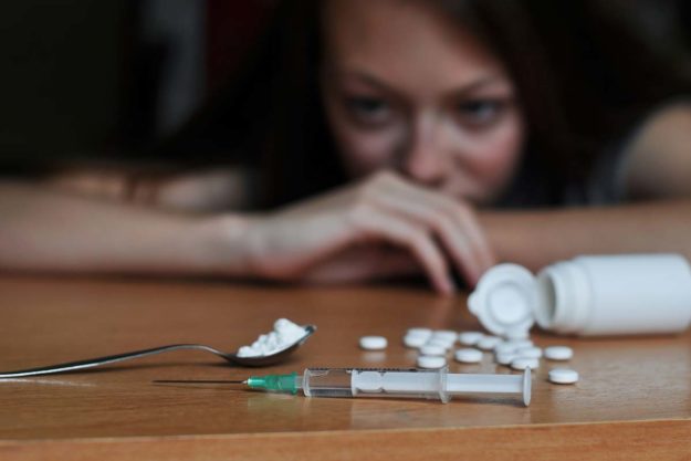 woman staring at pills and a needle on a table considering the symptoms of addiction