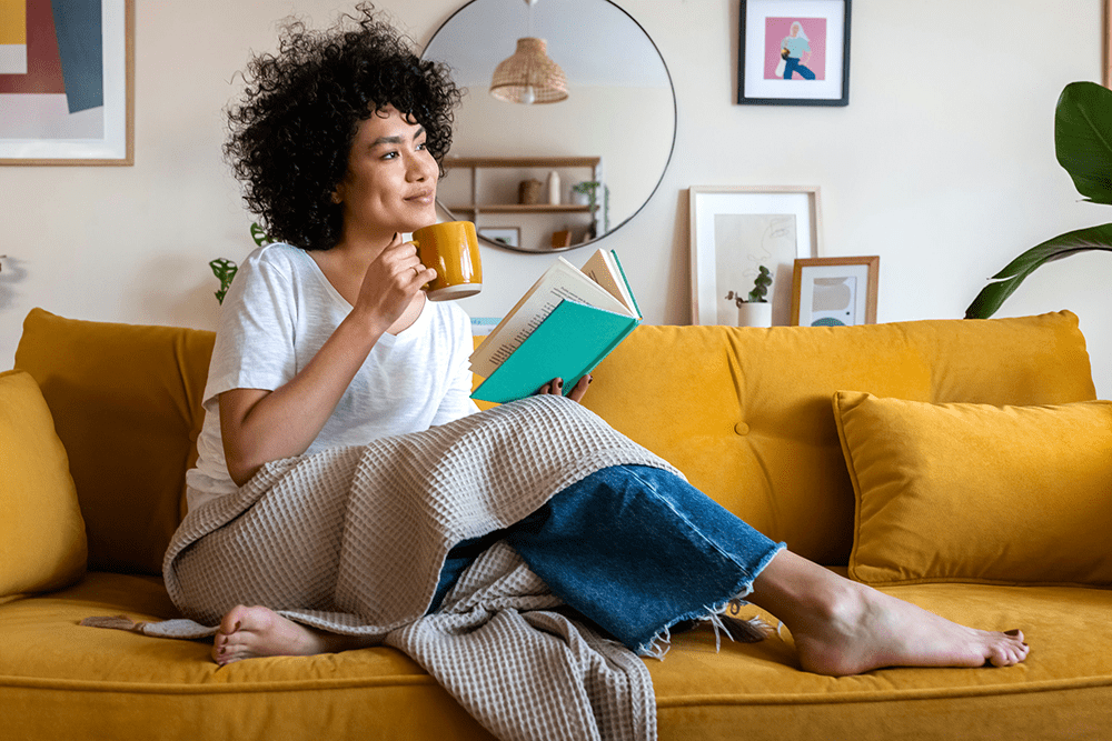 woman sitting on yellow couch with a book and a drink